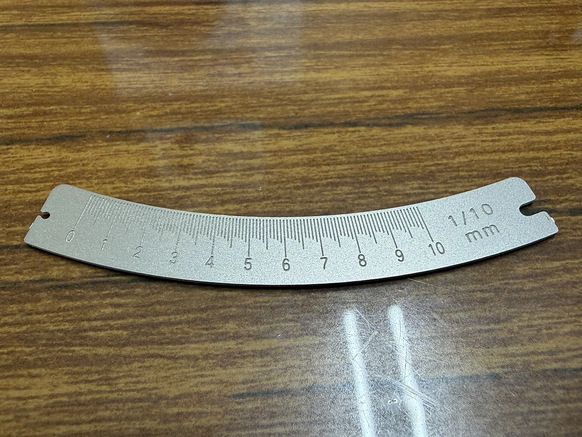 円弧状に湾曲したステンレス製の目盛りスケール。1/10mm刻みの目盛りがエッチングで入っている